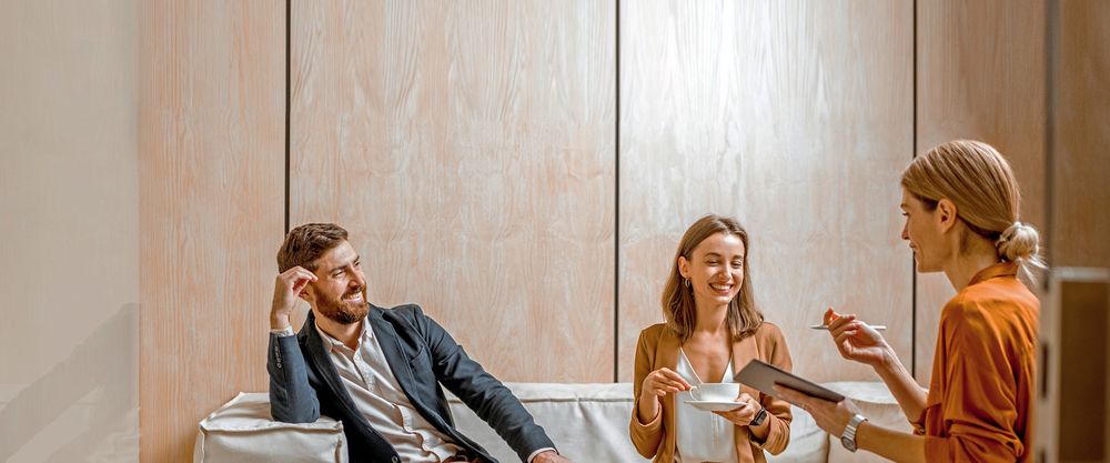 Ein fröhliches Paar sitzt auf einer Couch in einem minimalistischen Raum mit Holzverkleidung an der Wand und spricht mit einer Frau.